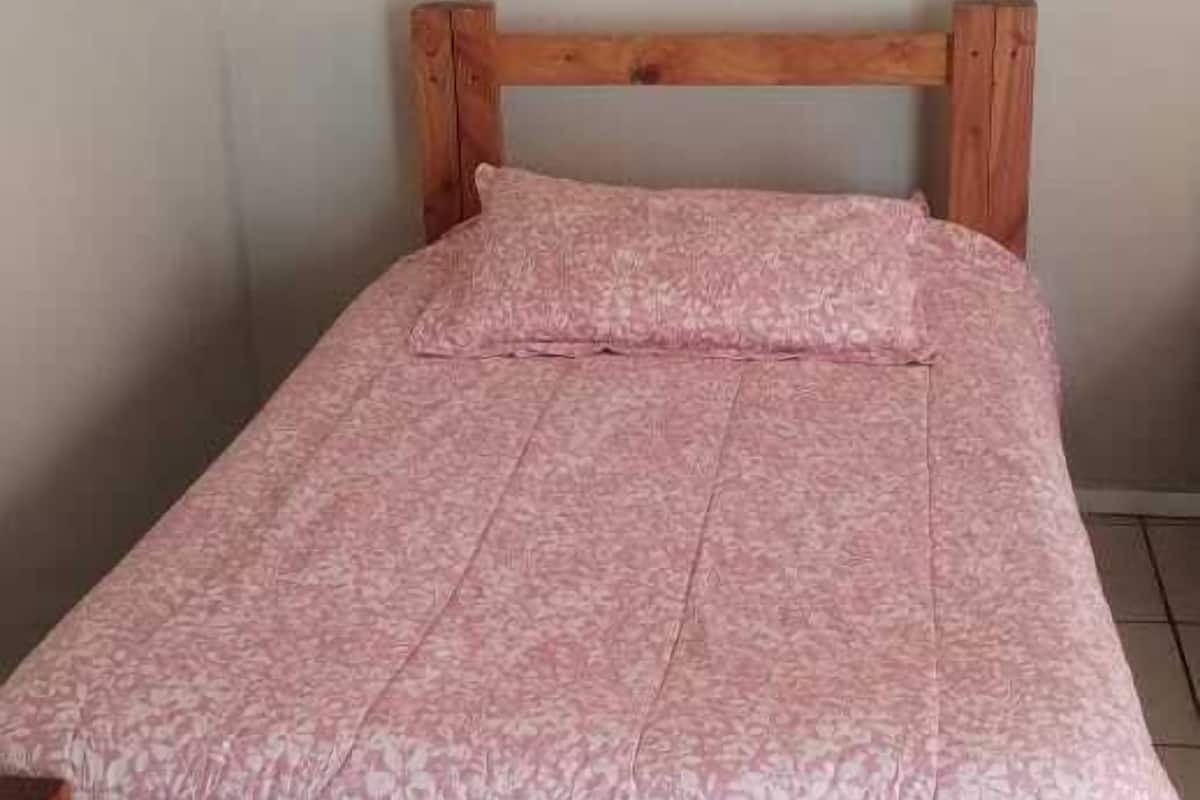 En säng i trä med ett rosa, mönstrat överkast i ett rum med ett klinkergolv och en inramad tavla på väggen.