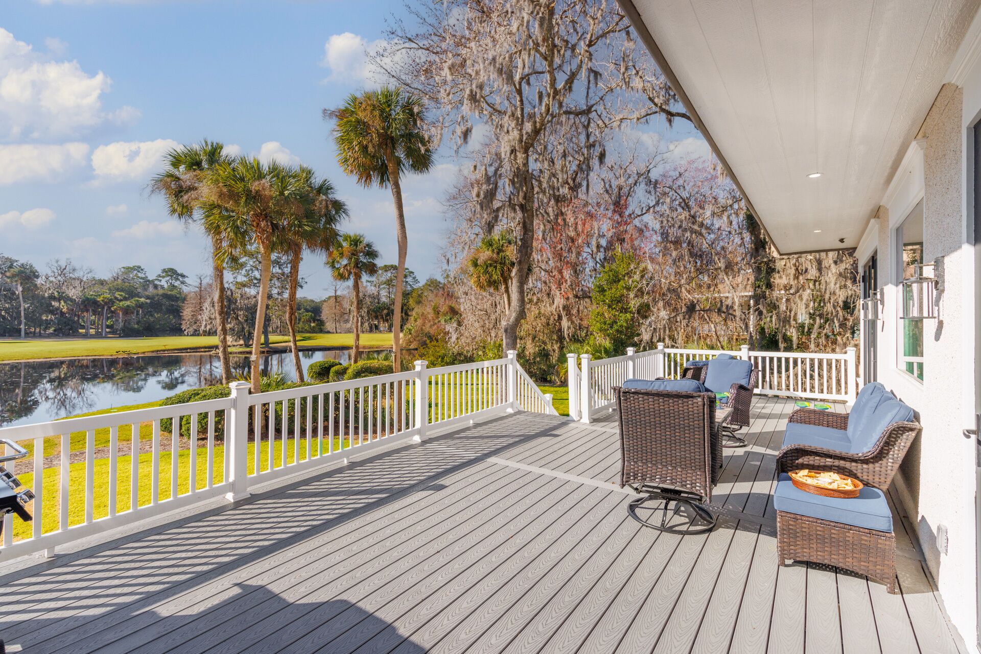 A covered deck with white railings, wicker furniture, and a view of a golf course and water.
