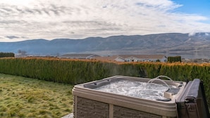 A hot tub with a stone exterior, surrounded by a well-maintained hedge, set against a backdrop of mountains and a lake.