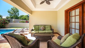 Un patio iluminado por el sol con mobiliario de mimbre, un ventilador de techo y vistas a una piscina.