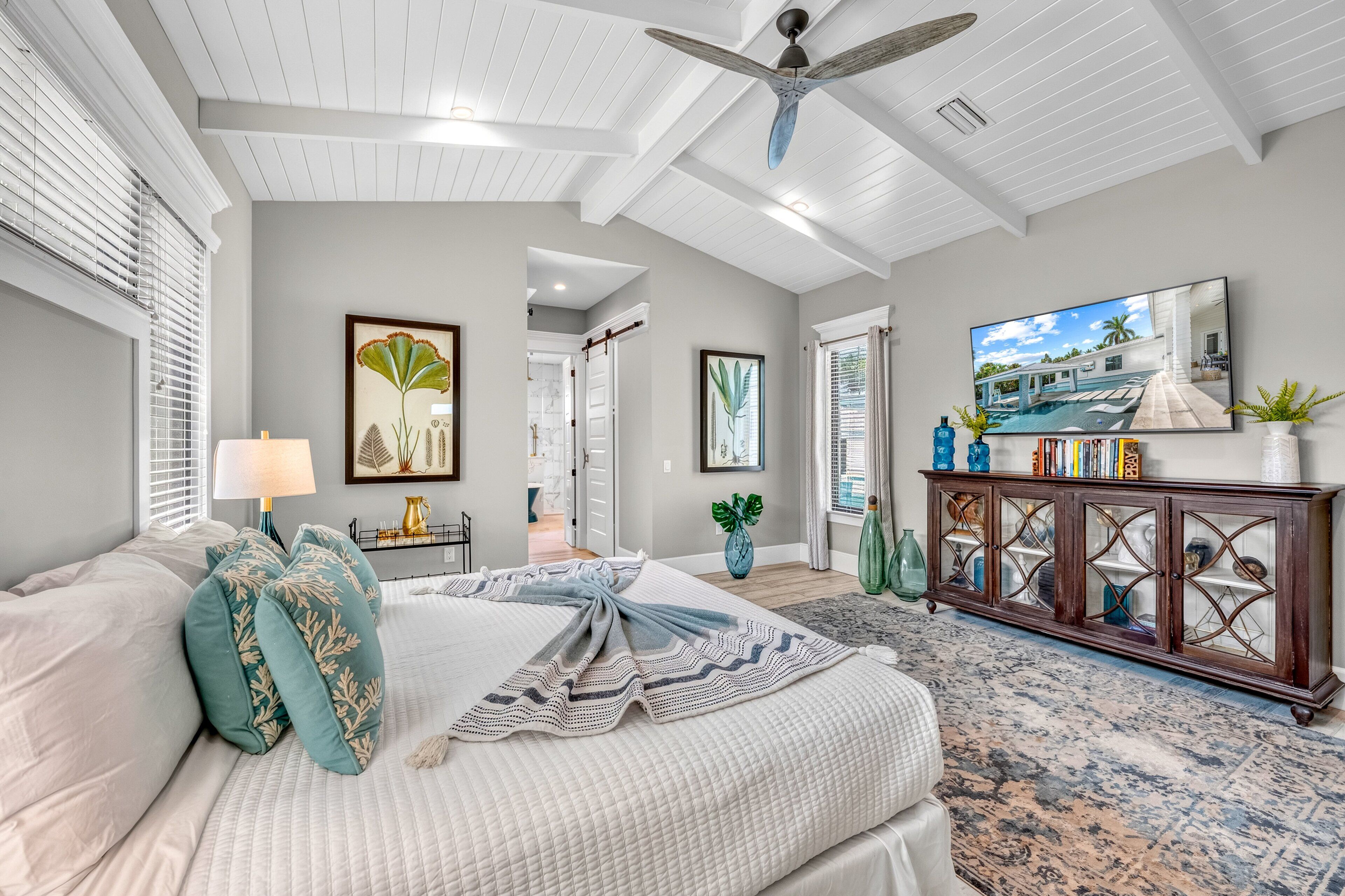 A bedroom with a bed, a ceiling fan, a large window with blinds, a television, and a cabinet with decorative items.