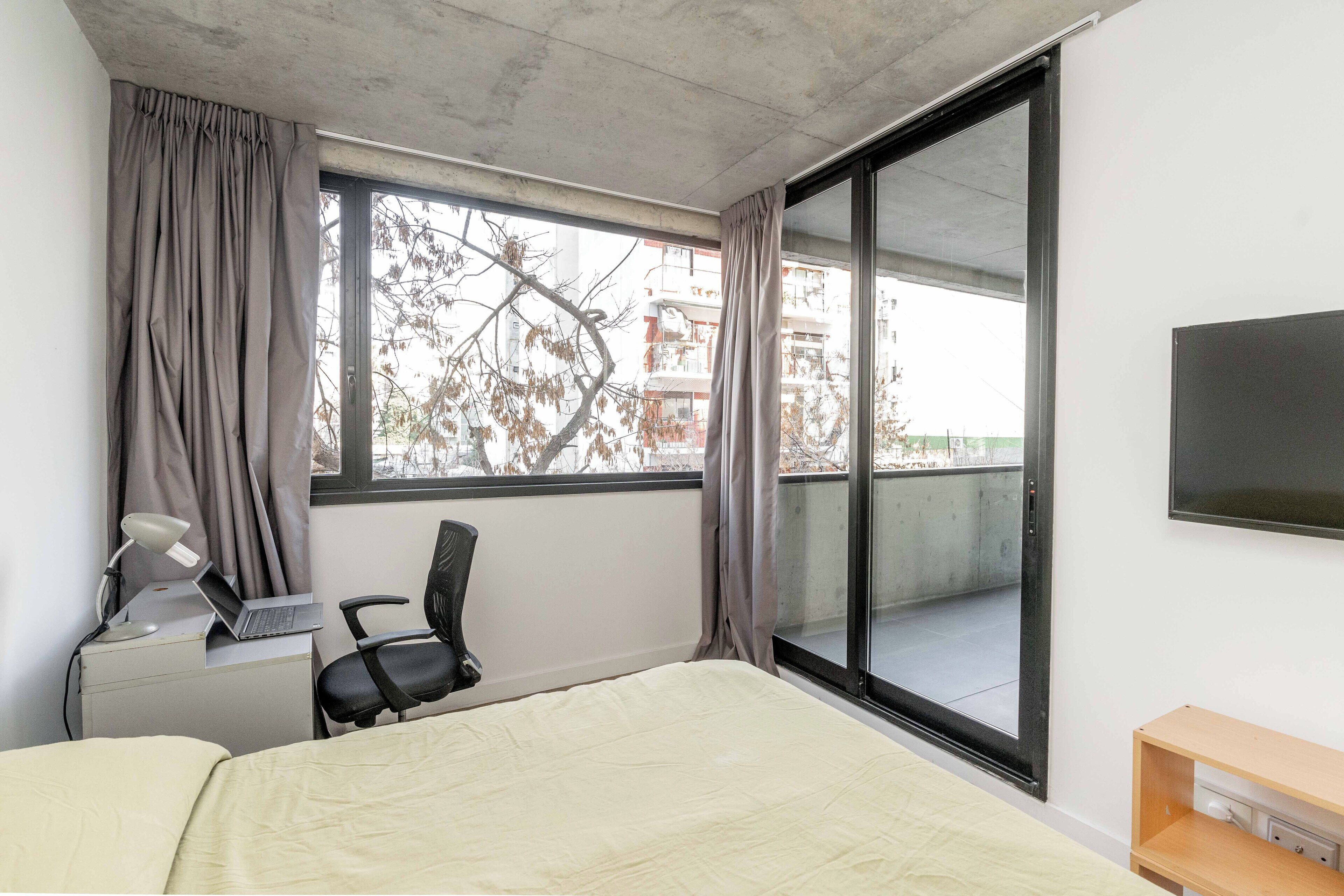 Una camera d'albergo con un letto, una scrivania con un portatile, una sedia, una televisione e un'ampia finestra con tende.