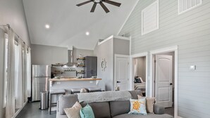 A modern living room with a gray sofa, a ceiling fan, and a kitchen in the background. - Schipps Creek - Ocean & Bay Views (Bolivar Peninsula)