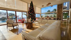 Un salon spacieux, agrémenté d’un grand sapin de Noël, de canapés en cuir blanc et offrant une vue sur l’extérieur grâce à de grandes fenêtres.