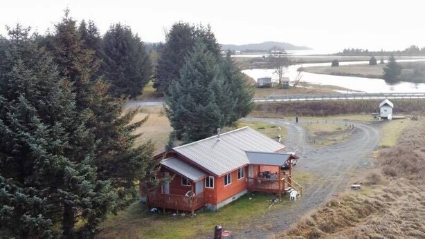 A cabin with a porch, surrounded by trees and a dirt path leading to a bridge over water.