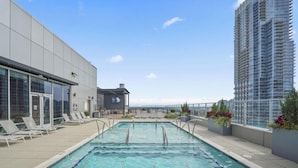 A rooftop pool with lounge chairs, a modern building, and a high-rise building in the background. - Reluxme | Lux 3BR Condo w/ Skyline View + Stadiums (Charlotte)