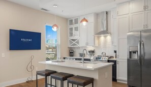 A modern kitchen with white cabinets, a stainless steel refrigerator, a central island with stools, pendant lighting, and a city view through the window.