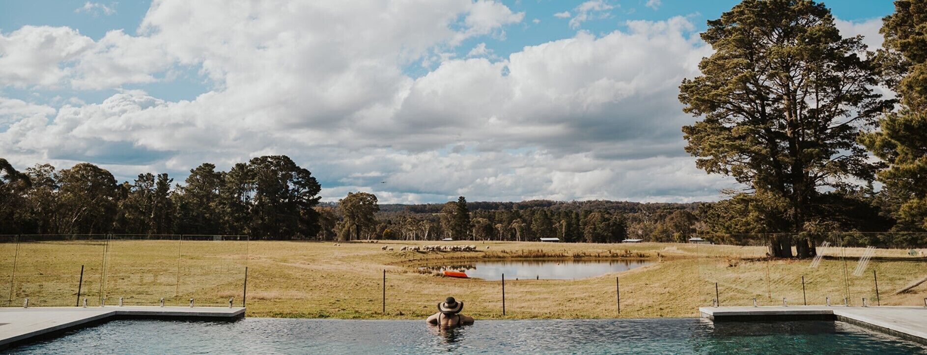 Une piscine avec une personne dans l’eau, entourée d’une zone délimitée par une clôture, avec des arbres et une étendue d’eau visible au loin.