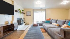 A modern living room with a grey sofa, a flat-screen TV, a wooden cabinet, and a large window. - Spacious 6 BR Extended Stay (North Salt Lake)