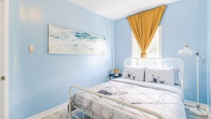Ein Schlafzimmer mit Bett, einem Fenster mit gelben Vorhängen, einem Nachttisch mit Lampe und einem Wandbild.