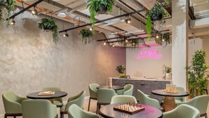 A modern interior with round tables and green chairs, a neon sign, and hanging plants.
