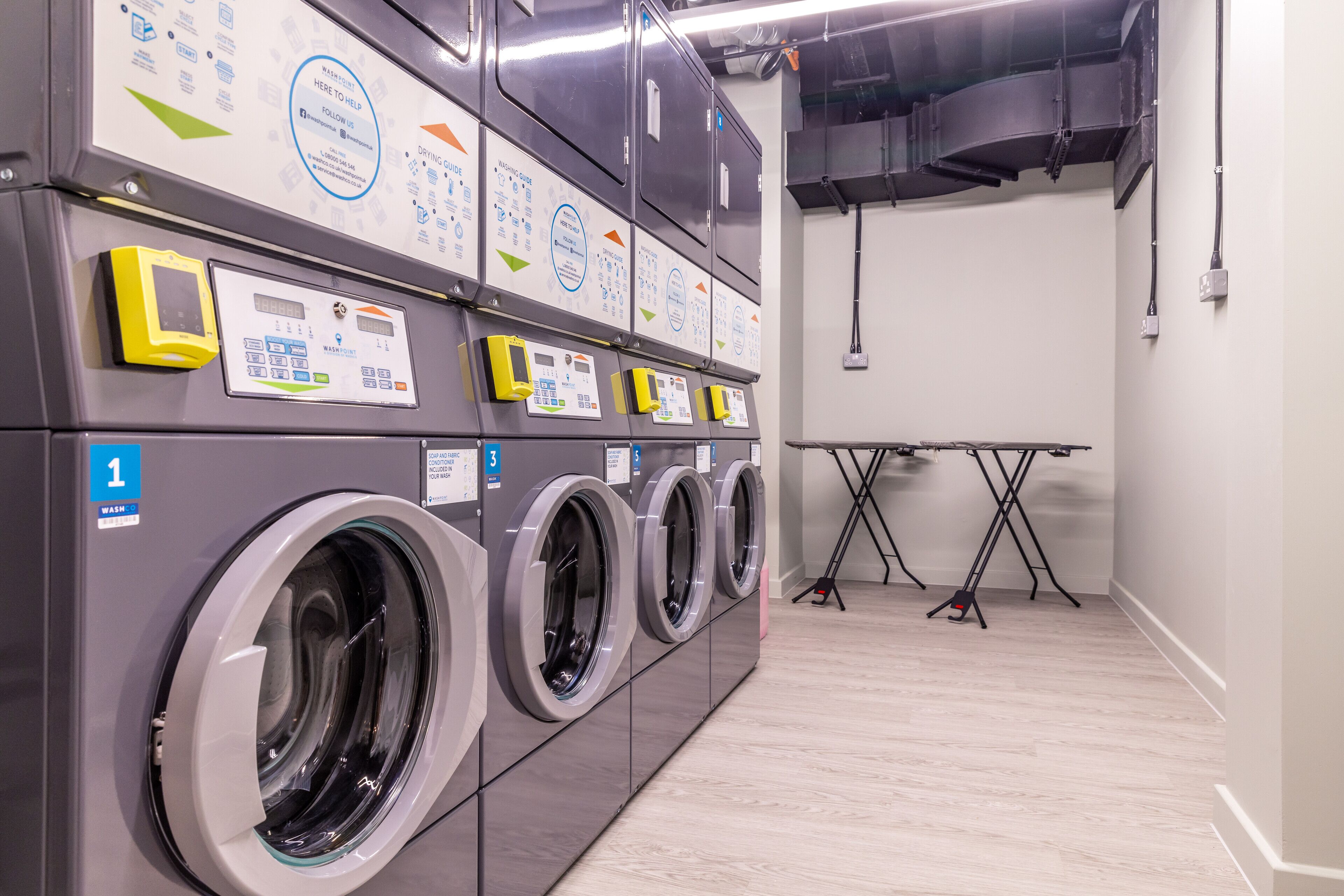 A row of front-loading washing machines with control panels, a folding table, and a power outlet on the wall.