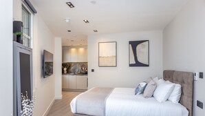 A modern bedroom with a large bed, a built-in TV, and a view into the kitchen.