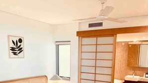 Ein Schlafzimmer mit Bett, Nachttisch, Lampe, Fenster und Deckenventilator, sowie ein Badezimmer mit Waschbecken und Spiegel.