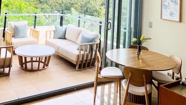 Ein modernes Wohnzimmer mit einer Couch, Sesseln, einem runden Tisch und einem Balkon mit Blick auf Grünflächen.