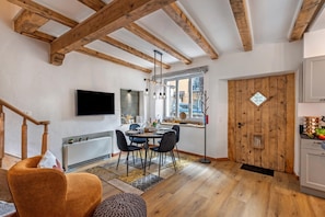 A modern kitchen with wooden beams, a dining area with a table and chairs, a TV mounted on the wall, and a wooden door with a window.