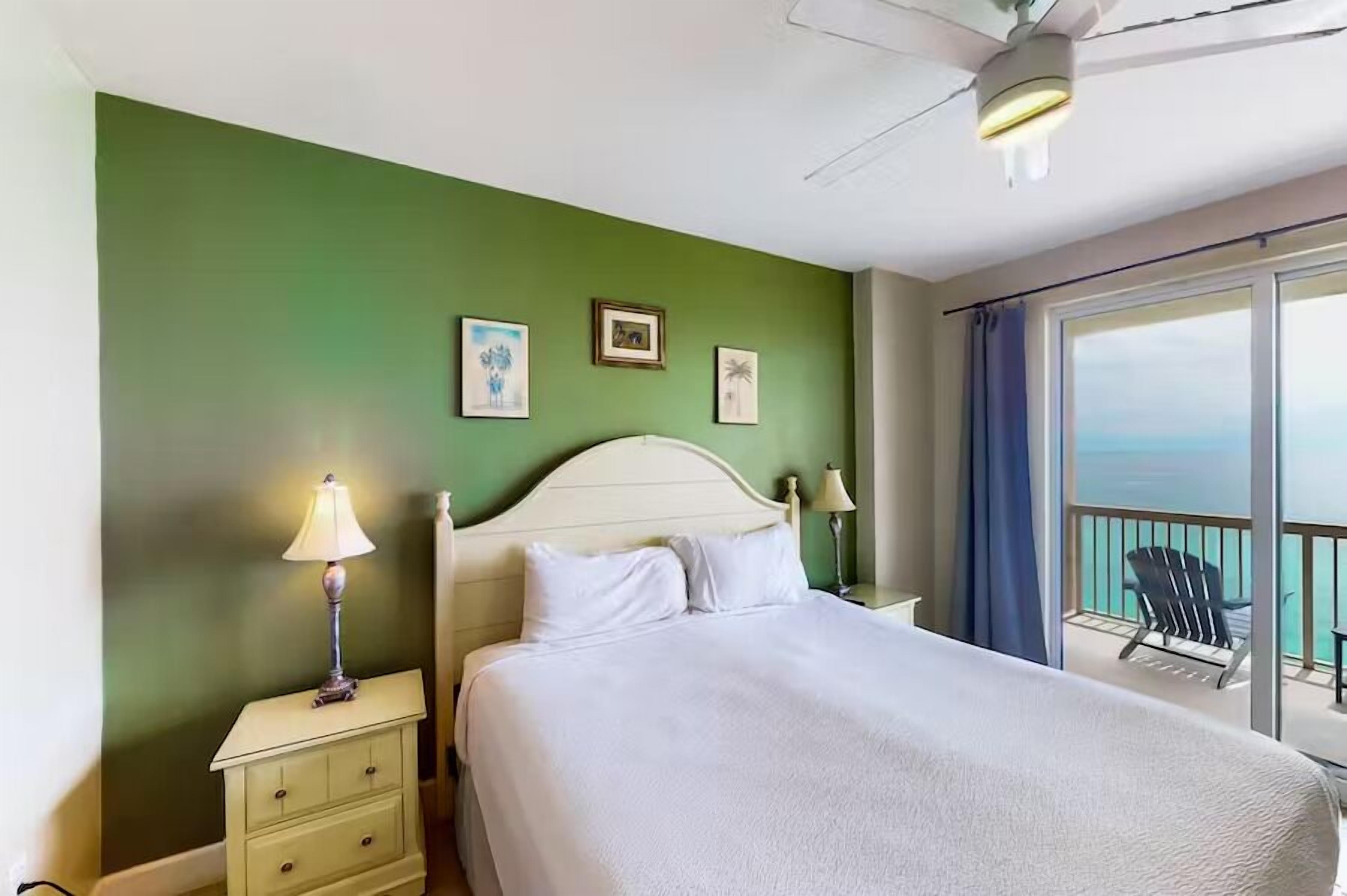 Camera d'albergo con un letto, un comodino, una lampada, una ventola a soffitto e un balcone con una sedia.