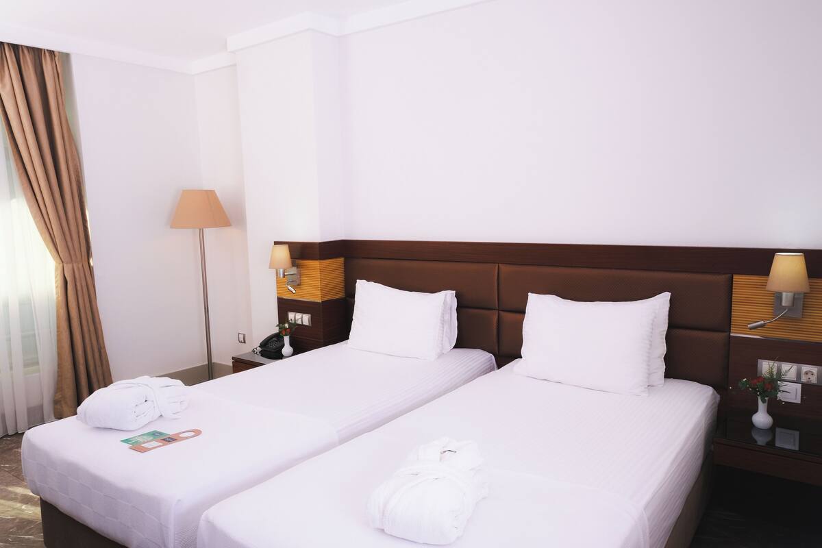 Habitación de hotel con dos camas, una mesita de noche, una lámpara y una ventana con cortinas.