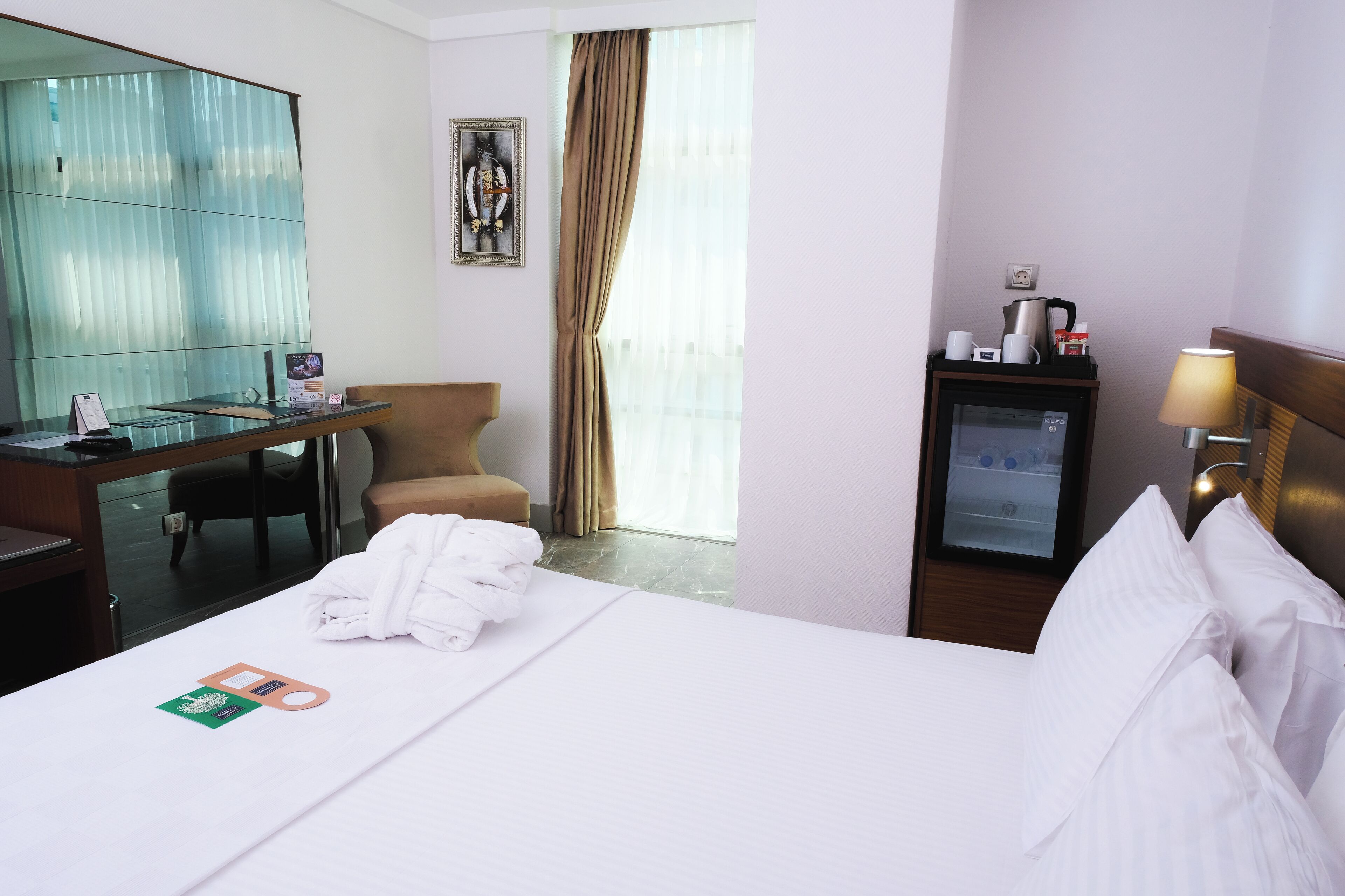 A hotel room with a bed, a desk, a chair, a minibar, and a window with curtains.