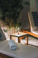 A room with a wooden chair, a small table with a lamp, and a rolled towel on a cushioned bench.