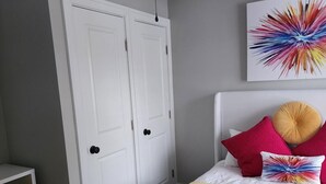 Une chambre à coucher avec un lit, un ventilateur de plafond, une porte, une chaise et une œuvre d’art colorée accrochée au mur.