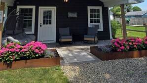 Un porche abrité avec un toit rouge, deux chaises, une petite table et des jardinières ornées de fleurs roses.