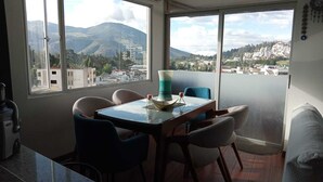 A dining area with a table and chairs, a view of a mountainous landscape, and a balcony with a railing. - Departamento con Jacuzzi 5 Piso el Condado (Quito)