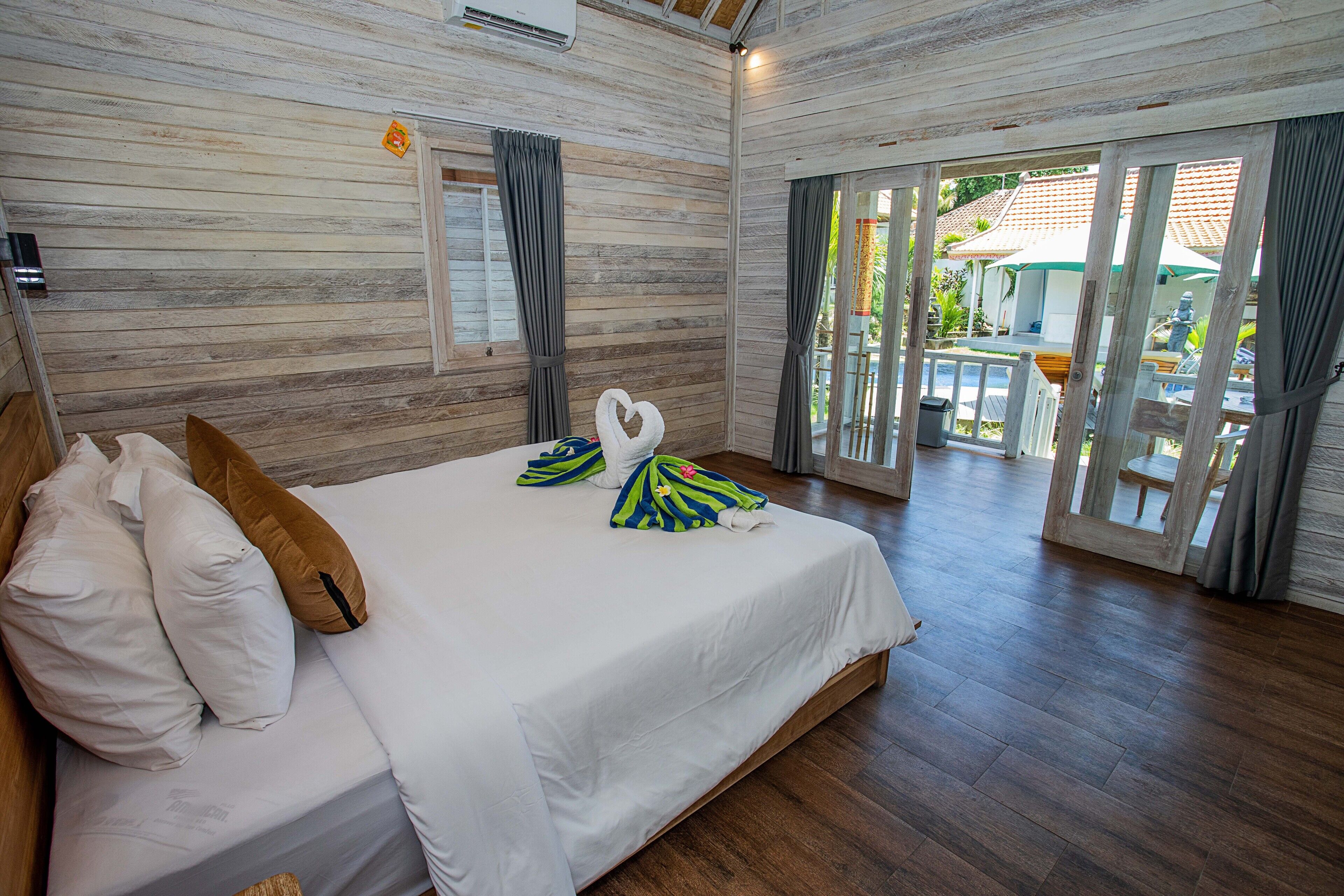Habitación de hotel con piso de madera, cama con ropa blanca y vista a un espacio exterior con una mesa y sillas a través de un vidrio.