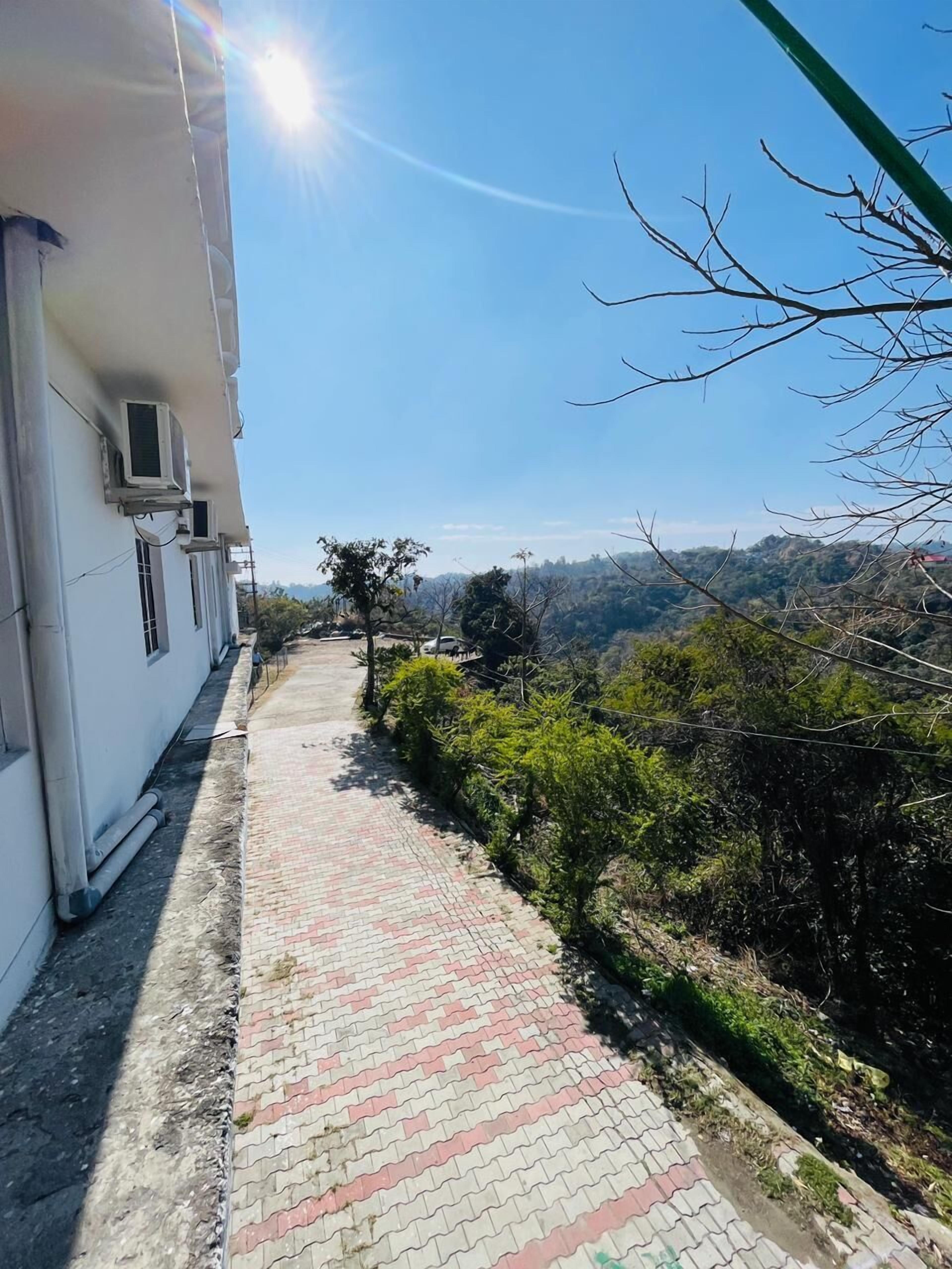 A paved pathway beside a building with air conditioning units, leading to a hilly landscape.