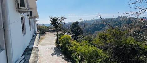 A paved pathway beside a building with air conditioning units, leading to a hilly landscape.