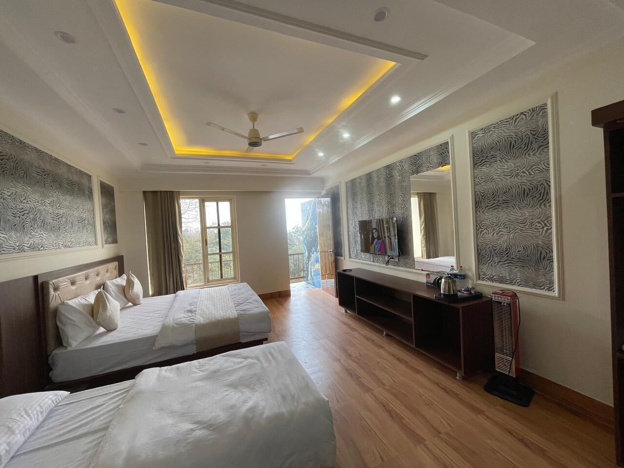 Habitación de hotel con dos camas, televisión, ventilador de techo y un ventanal grande.