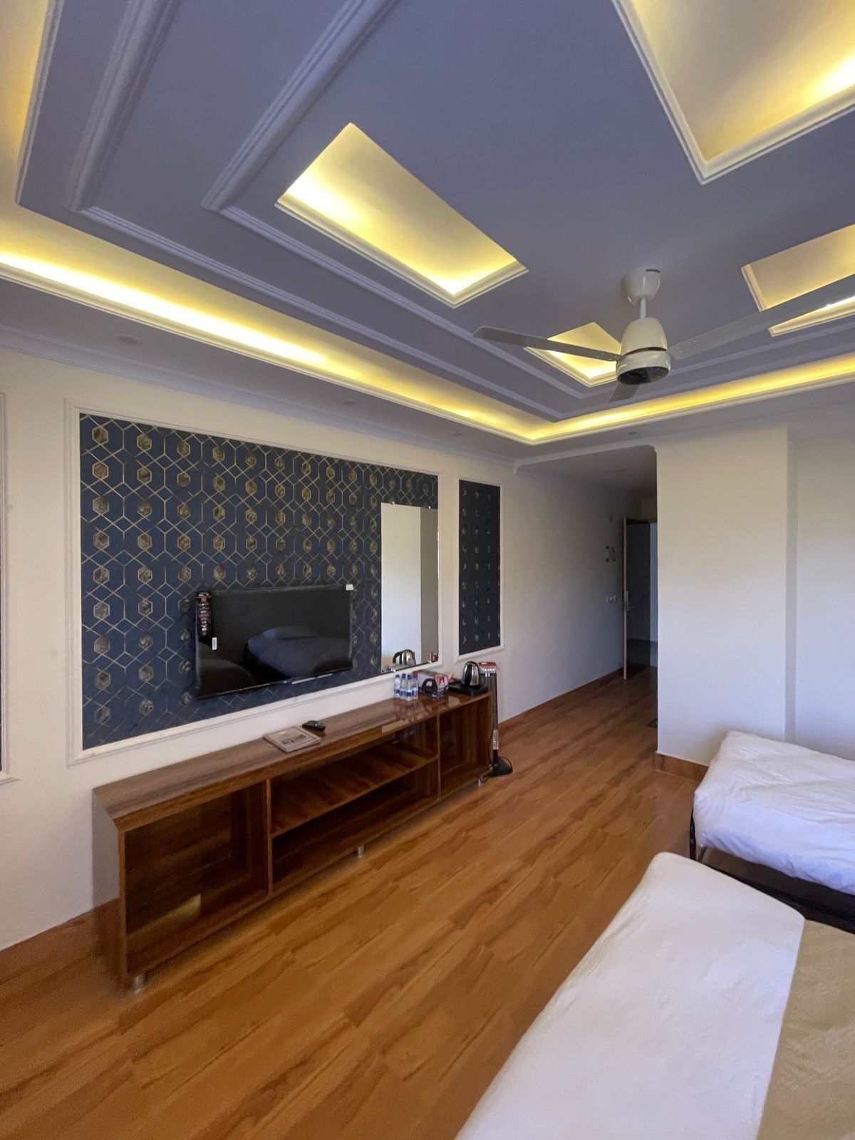 A hotel room with a wooden floor, a TV mounted on the wall, a ceiling fan, and a bench.