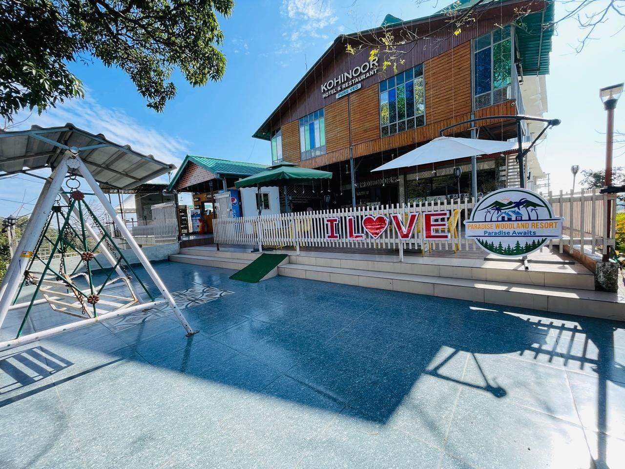Un hôtel avec une enseigne indiquant « Kohinoor Hotel & Restaurant », un ensemble de balançoires, un espace piscine et une autre enseigne sur laquelle on peut lire « J’adore le complexe Paradise Woodland ».