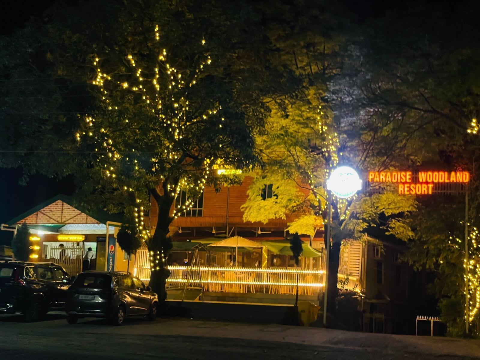 Un edificio con un letrero que dice 'Paradise Woodland Resort' por la noche, rodeado de árboles y automóviles estacionados.