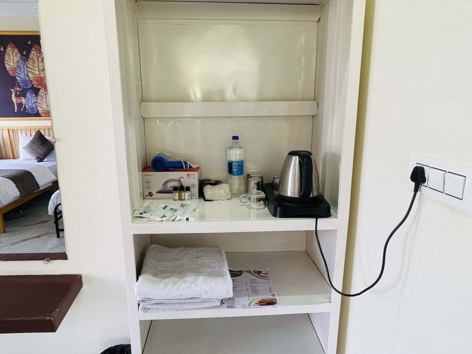 Una habitación de hotel con una cama, estante con artículos de aseo, hervidor y toma de corriente.
