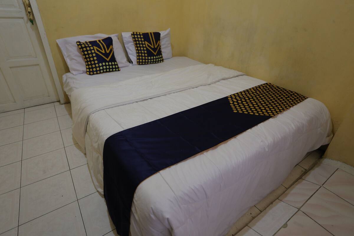 Una cama con ropa de cama blanca y una manta azul oscuro, dos almohadas con diseños amarillos y negros, y una pared amarilla lisa.