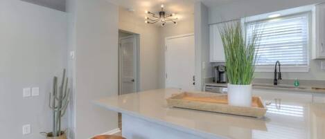 A modern kitchen with a central island, bar stools, a hanging light fixture, and a potted plant.