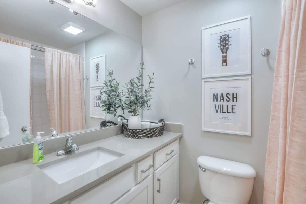Un baño moderno con lavamanos, espejo y retrete blancos. Hay cuadros enmarcados en la pared y una planta en maceta sobre el mueble.