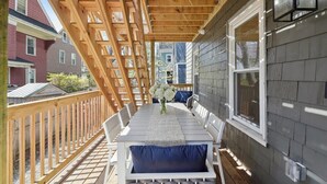 Een overdekt terras buiten met een houten terras, witte meubels en uitzicht op omliggende huizen.