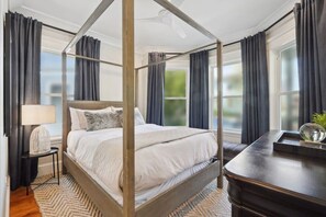 A four-poster bed with a wooden frame, a bedside table with a lamp, and a view of the outdoors through large windows with dark curtains. - 6BR Classic Boston Home | Near Savin Hill T (Boston)