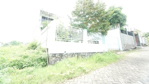 A building with a gated entrance, a paved pathway, and a tree in the background. - Hotel O Kencana Homestay Syariah Near Masjid AL-BAROKAH Kanyuran (Mojokerto)