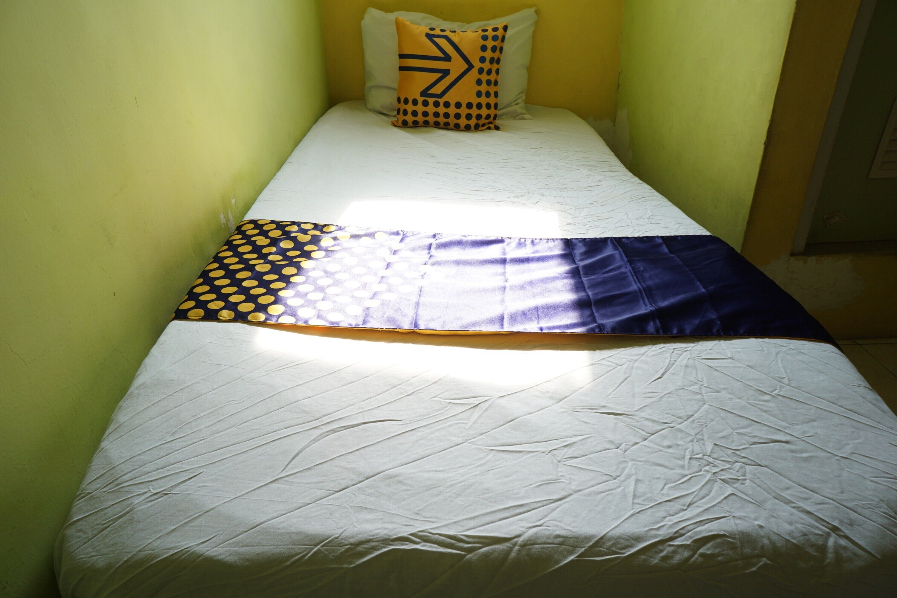 Una habitación pequeña con cama individual, colcha blanca, manta con estampado azul y amarillo, y almohada con diseño geométrico.