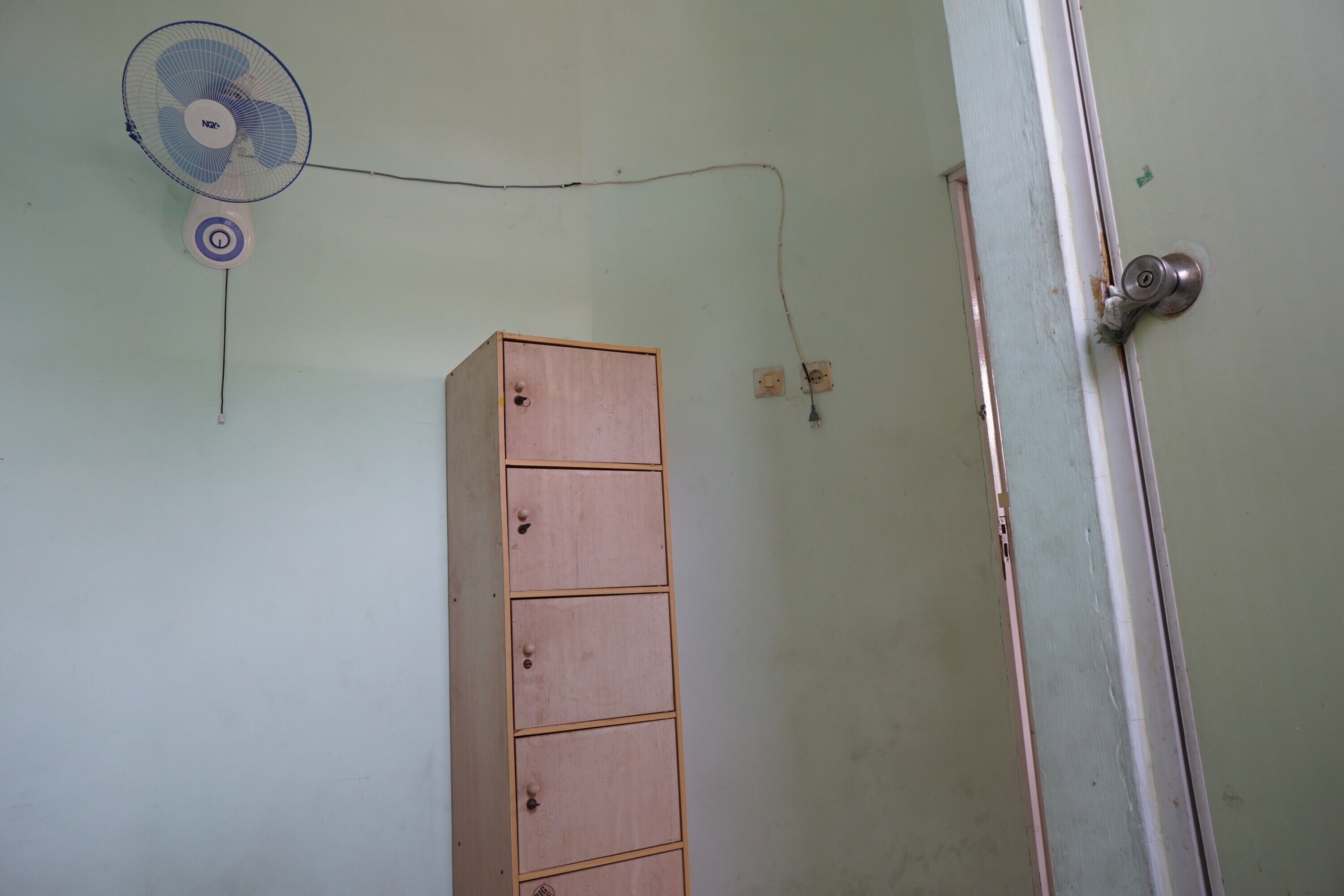 Une pièce avec un meuble en bois, un ventilateur fixé au mur et une porte dotée d’une poignée en métal.