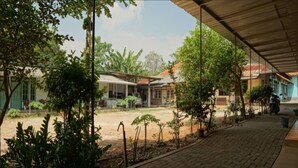 A covered outdoor area with potted plants, a paved walkway, and buildings in the background. - SPOT ON 93611 Kost Pak Suyatno Syariah 2 (Ponorogo)