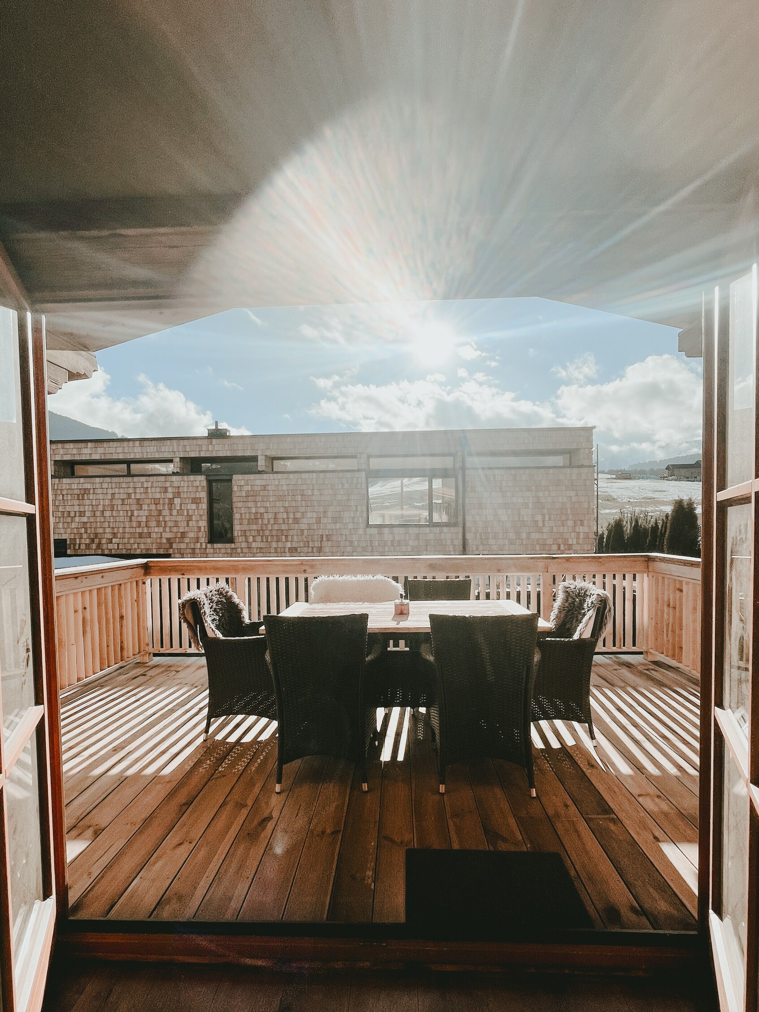 Eine Holzterrasse mit Tisch und Stühlen, Blick auf ein Gebäude und ein klarer Himmel.