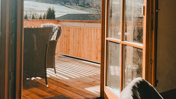 A room with a wooden floor, a wicker chair, and a view of a snowy landscape through a glass door. - Apartment 'Katie' with Mountain View, Private Terrace and Wi-Fi (Hopfgarten)