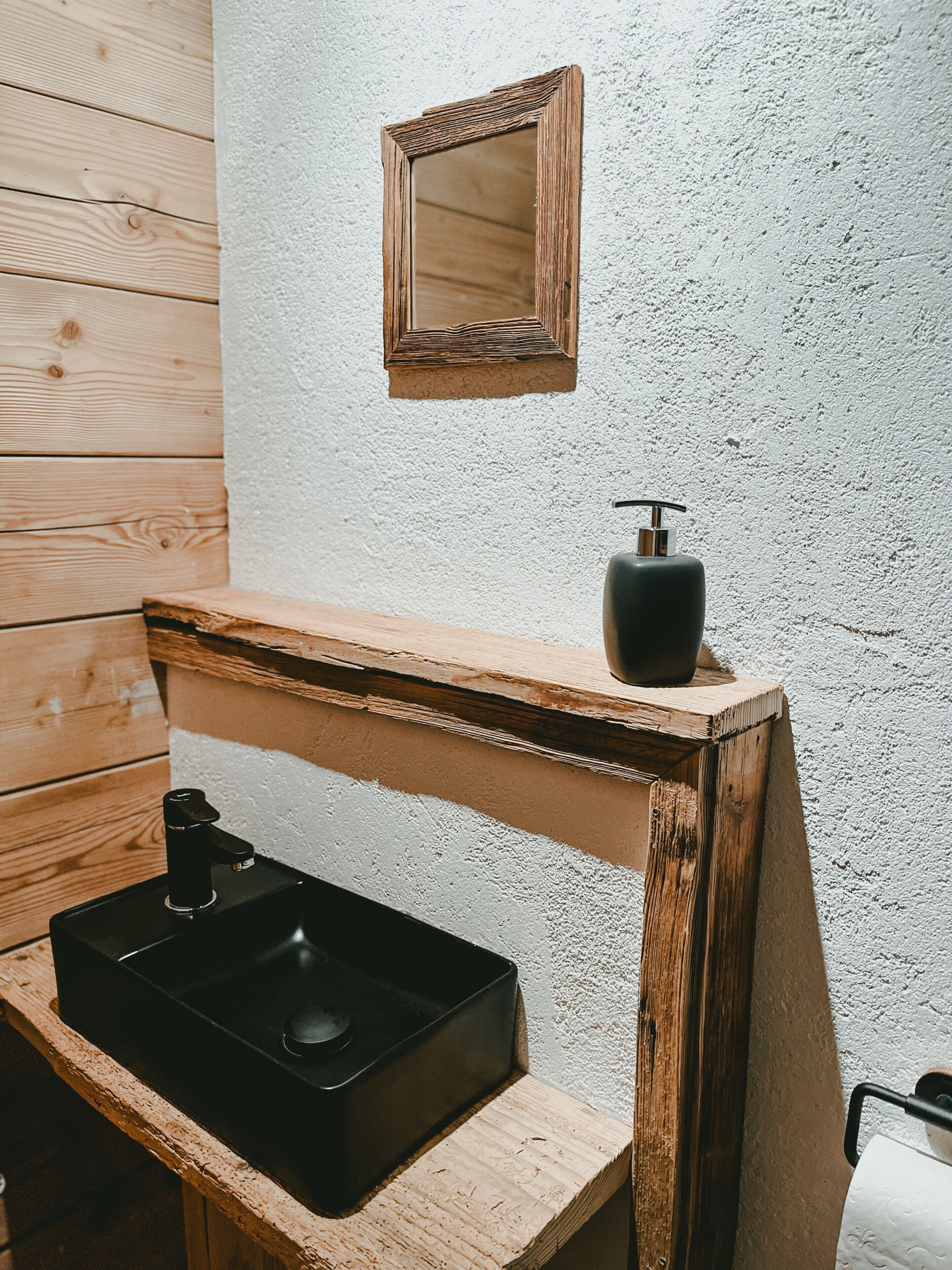 Un bagno con un mobile bagno in legno, un lavandino nero, uno specchio e un dispenser di sapone.