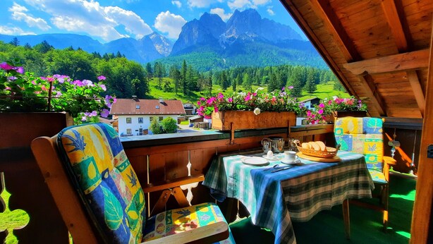 Una cabaña alpina con una mesa lista para el desayuno, muebles de madera y vistas a montañas y vegetación.