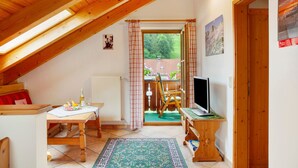 Una habitación acogedora con techo inclinado, una mesa de madera con una botella y vasos, un televisor sobre un mueble, y vistas a un espacio exterior a través de una puerta.