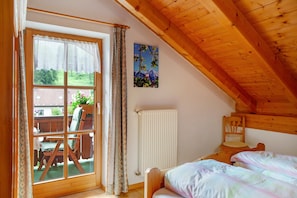 Une chambre avec un plafond en bois, un lit recouvert d’une couverture colorée, une chaise, un radiateur et une fenêtre avec des rideaux.